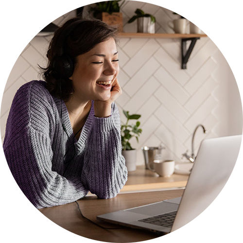 A woman in a knit jumper sits at her kitchen counter looking at a silver laptop and smiling