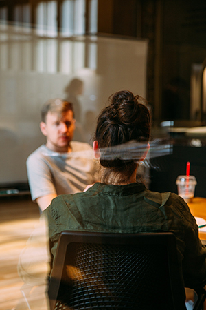 A casually dressed man sits at a desk opposite a woman in business attire inside a glass office.