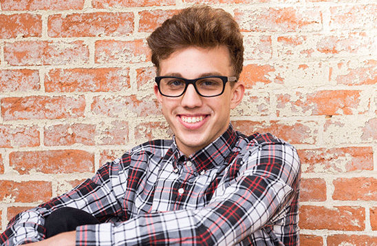 Young man wearing glasses and a check shirt sitting against a wall and smiling